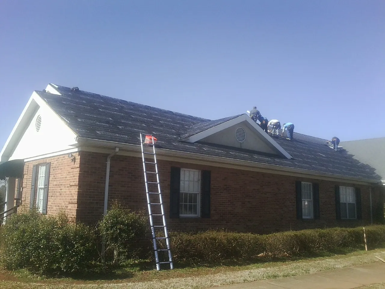 Crew tearing shingles off a brick ranch roof with a ladder set — Metal Roof Repair work by our crew in Lost Nation