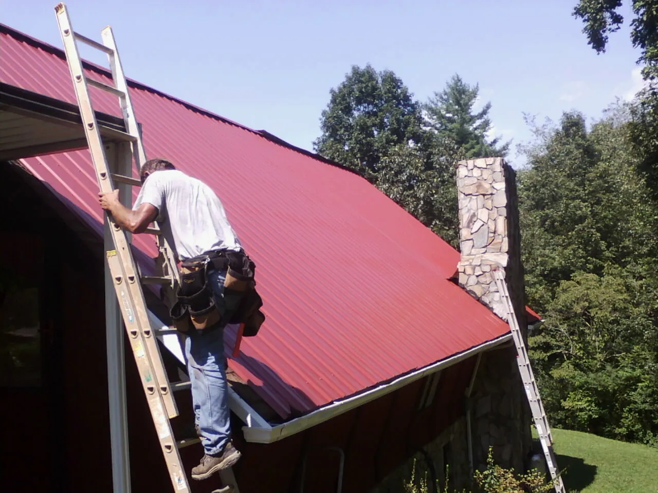 Roofer on a ladder beside a red metal roof and stone chimney — Soffit & Fascia Repair by Roofing Brothers in Lost Nation